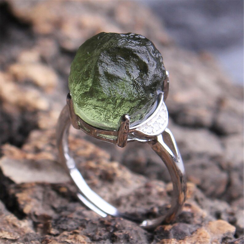 May include: A silver ring with a large, green, rough-cut gemstone. The ring has a delicate, curved band with small, clear stones.