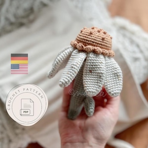Könnte beinhalten: Eine gehäkelte Gänseblümchen mit braunem Zentrum und hellgrauen Blütenblättern. Das Gänseblümchen wird in einer Hand gehalten. Ein PDF-Häkelmuster wird im Bild beworben. Das Bild enthält auch die Flaggen Deutschlands und der Vereinigten Staaten.