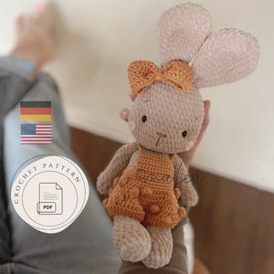 Könnte beinhalten: Eine Häkelanleitung als PDF mit einem hellbraunen Plüschhasen mit langen Ohren, einer kleinen orangefarbenen Schleife am Kopf und einer orangefarbenen, strukturierten Latzhose mit Noppenmuster. Das Bild enthält deutsche und amerikanische Flaggen.