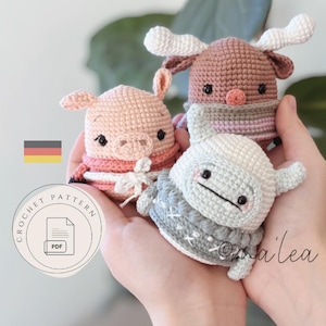 Könnte beinhalten: Drei gehäkelte Amigurumi-Tiere: ein Schwein, ein Rentier und ein Wal. Das Schwein ist rosa, das Rentier ist braun und der Wal ist grau. Jedes hat einen Schal. Das Bild enthält ein PDF-Häkelmuster-Logo und eine deutsche Flagge.