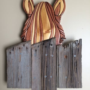 May include: A wooden horse sculpture peeking over a weathered wooden fence. The horse is carved with intricate details and has a reddish-brown and light brown color scheme. The fence is made of reclaimed wood planks with visible screw heads.
