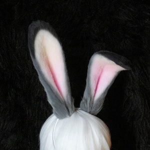 Puede incluir: Unas orejas de conejo grises y blancas con interior rosa, sujetas a una diadema blanca, y una cola a juego. Los artículos se muestran sobre una cabeza de maniquí blanca, sobre un fondo negro.