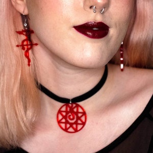 Peut inclure: Une personne aux cheveux roses portant un haut en maille noire avec un collier ras du cou rouge orné d'un pendentif en forme d'étoile. Le pendentif a un motif en spirale au centre. La personne porte également des boucles d'oreilles rouges avec un design en forme d'étoile similaire.
