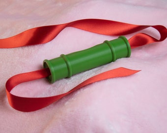 May include: A green cylindrical object wrapped in a red ribbon on a pink fuzzy surface.