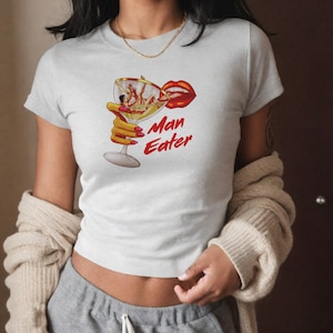 May include: White t-shirt featuring a graphic of a hand holding a martini glass with a couple inside. The text "Man Eater" is printed below the glass.
