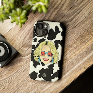Op de afbeelding: Een zwart-witte telefoonhoes met een koe-print met een kleurrijke illustratie van een vrouw met een zonnebril. De hoes heeft een zwarte cirkel met de tekst "OPA" in wit.