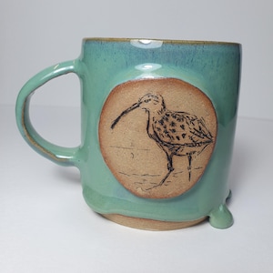 May include: A ceramic mug with a glossy teal glaze and a single handle. The front features a circular, unglazed tan medallion with a dark, etched line drawing of a long-billed shorebird standing on the ground. The mug has small, rounded feet.
