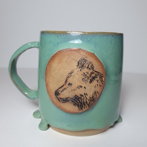 Op de afbeelding: Een keramische mok met een glanzend blauwgroen glazuur en een glad, lichtbruin rond medaillon aan de voorzijde. Het medaillon bevat een gedetailleerde zwarte inktillustratie van een berenkop in profiel. De mok heeft een gebogen handvat en voetjes.