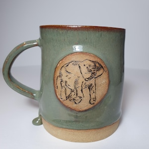 May include: A sage green ceramic mug with a brown rim and base, featuring a circular tan medallion on the front. The medallion displays a dark, etched line drawing of an elephant. The mug has a curved handle and a smooth, glazed finish.