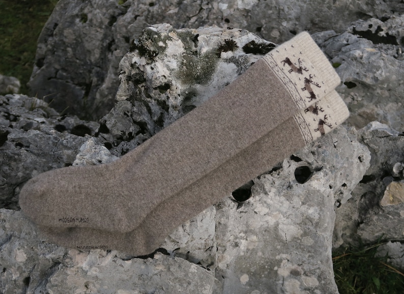 Puede incluir: Un par de calcetines de lana marr&oacute;n hasta la rodilla con un pu&ntilde;o blanco con un patr&oacute;n de renos. Los calcetines est&aacute;n doblados y colocados sobre una roca gris.
