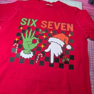 May include: A red t-shirt with the words "SIX SEVEN" in green and orange. The shirt features a graphic of a green hand making an "okay" gesture and a white hand wearing a Santa hat, with a black and white checkered background.