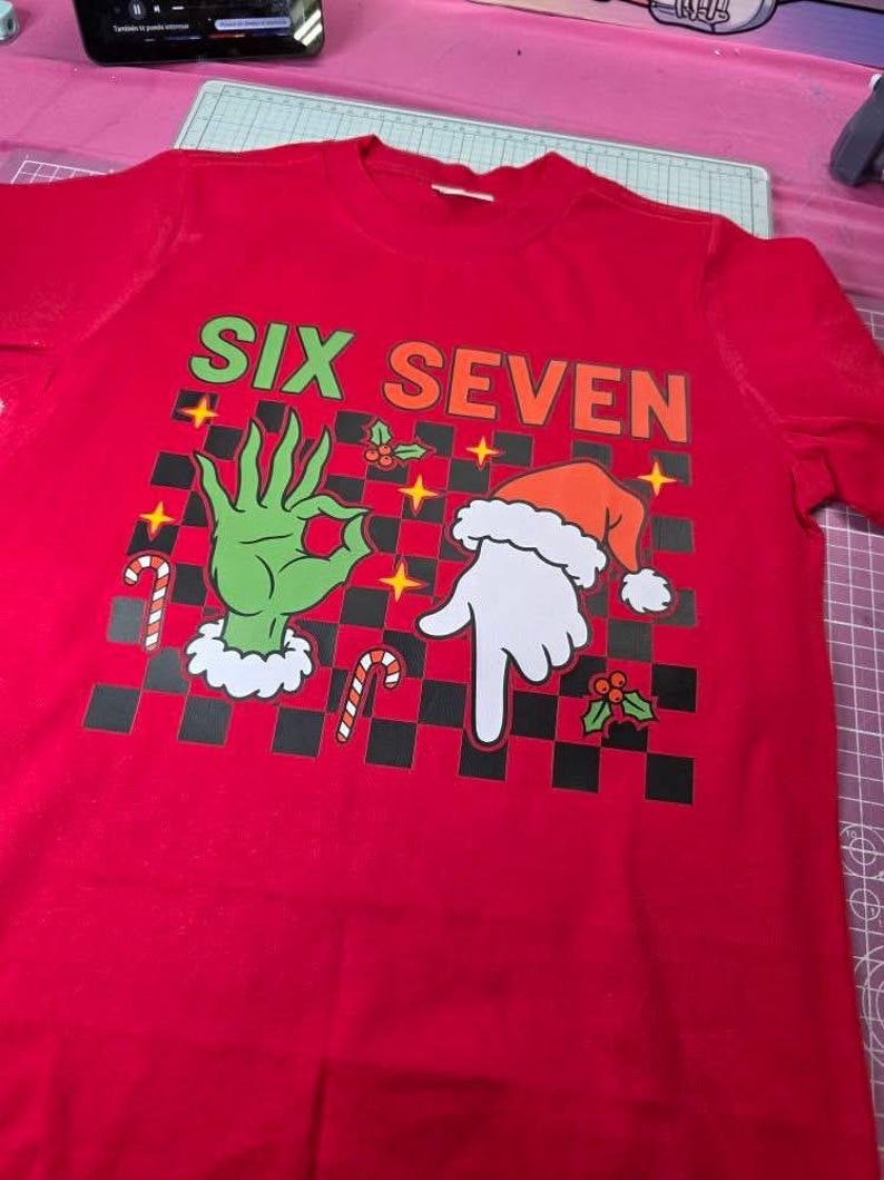 May include: A red t-shirt with the words "SIX SEVEN" in green and orange. The shirt features a graphic of a green hand making an "okay" gesture and a white hand wearing a Santa hat, with a black and white checkered background.