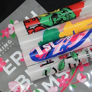 May include: Rolled vinyl sheets with colorful graphics, including a skull, abstract shapes, and floral designs. The sheets are stacked on top of a gray sheet with floral and text elements. The designs feature red, green, blue, and pink colors.