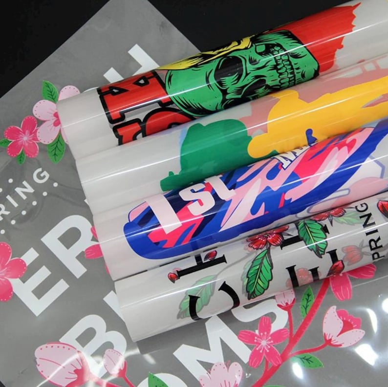 May include: Rolled vinyl sheets with colorful graphics, including a skull, abstract shapes, and floral designs. The sheets are stacked on top of a gray sheet with floral and text elements. The designs feature red, green, blue, and pink colors.