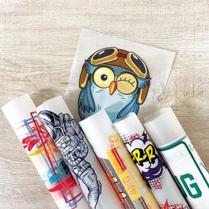 May include: A collection of rolled-up sticker sheets featuring various designs, including an astronaut, a cartoon owl wearing goggles, and comic book-style graphics. The stickers are on a light wood-grain surface.