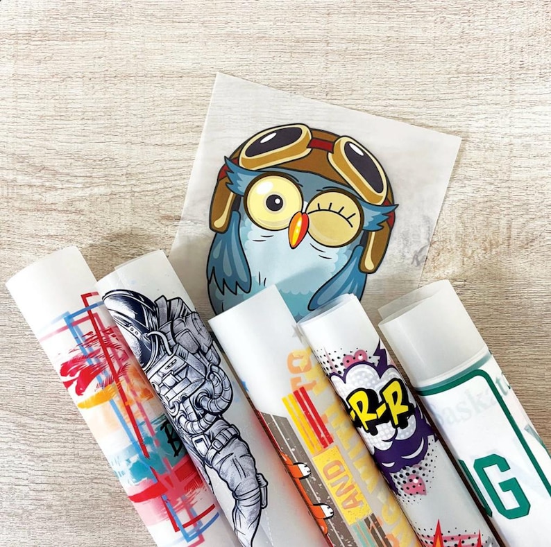 May include: A collection of rolled-up sticker sheets featuring various designs, including an astronaut, a cartoon owl wearing goggles, and comic book-style graphics. The stickers are on a light wood-grain surface.