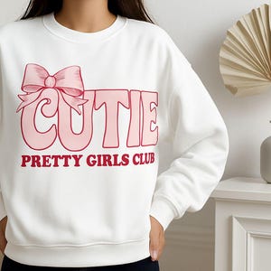 May include: White sweatshirt with the word "CUTIE" in pink block letters and a pink bow graphic. Below the word "CUTIE" is the text "PRETTY GIRLS CLUB" in red letters. The sweatshirt is a casual style.
