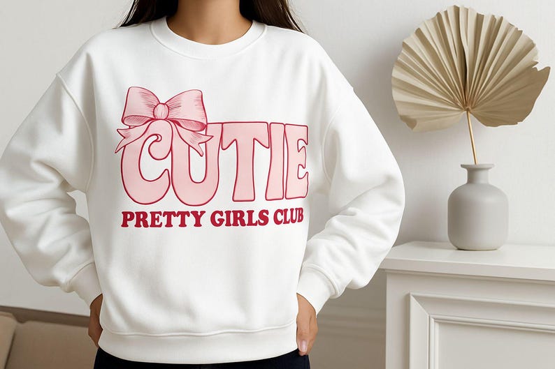 May include: White sweatshirt with the word "CUTIE" in pink block letters and a pink bow graphic. Below the word "CUTIE" is the text "PRETTY GIRLS CLUB" in red letters. The sweatshirt is a casual style.