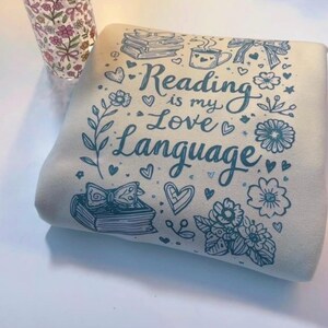 May include: A folded, cream-colored sweatshirt with a teal graphic that reads "Reading is my Love Language." The design includes illustrations of books, hearts, flowers, and a coffee cup. A floral patterned glass is visible in the background.