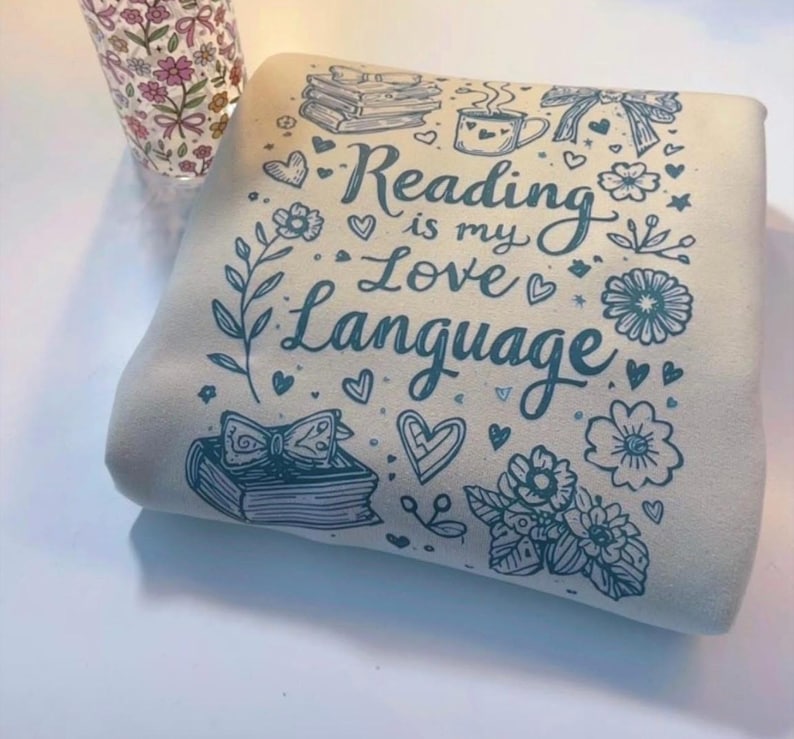 May include: A folded, cream-colored sweatshirt with a teal graphic that reads "Reading is my Love Language." The design includes illustrations of books, hearts, flowers, and a coffee cup. A floral patterned glass is visible in the background.