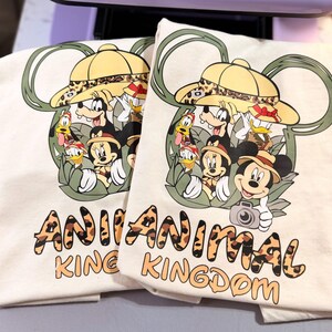 May include: Two off-white t-shirts with a colorful graphic featuring Mickey Mouse, Minnie Mouse, Donald Duck, and Goofy in safari hats. The text "ANIMAL KINGDOM" is printed in a leopard print font below the graphic.