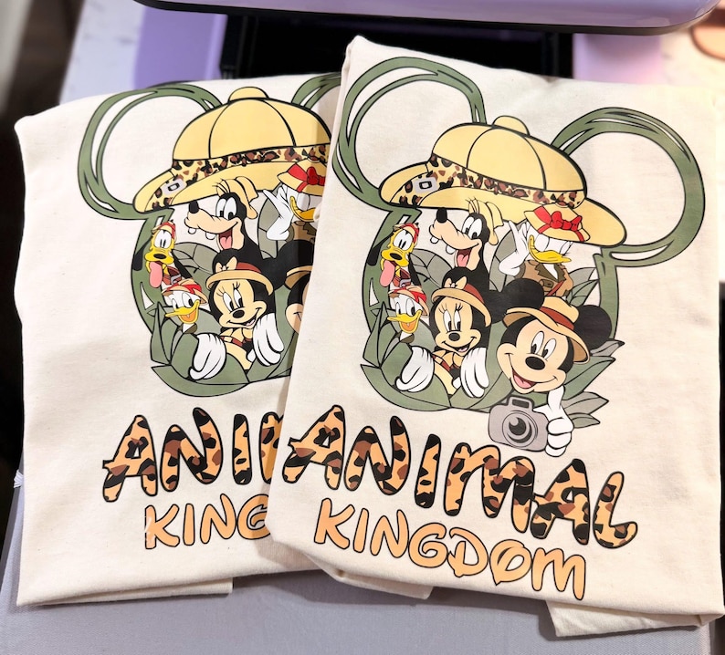 May include: Two off-white t-shirts with a colorful graphic featuring Mickey Mouse, Minnie Mouse, Donald Duck, and Goofy in safari hats. The text "ANIMAL KINGDOM" is printed in a leopard print font below the graphic.