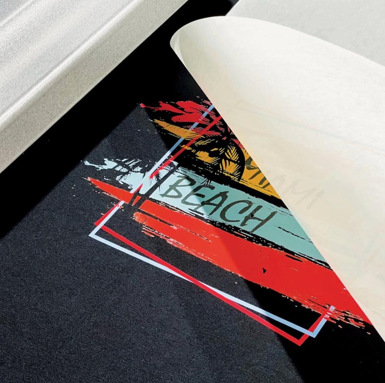 May include: Black fabric with a colorful beach-themed graphic. The design includes the word "BEACH" in white, set against red, blue, and yellow brushstrokes, with a palm tree silhouette. A white paper corner is peeling back.