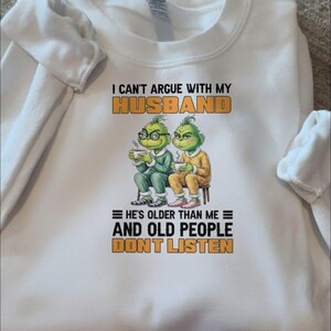 May include: White sweatshirt with the text "I CAN'T ARGUE WITH MY HUSBAND" in orange and black. Below the text are two cartoon Grinch characters, and the text "HE'S OLDER THAN ME AND OLD PEOPLE DON'T LISTEN".