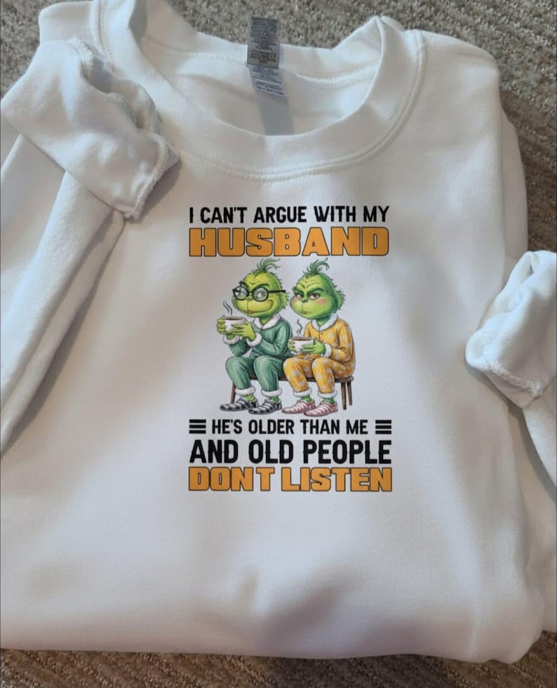 May include: White sweatshirt with the text "I CAN'T ARGUE WITH MY HUSBAND" in orange and black. Below the text are two cartoon Grinch characters, and the text "HE'S OLDER THAN ME AND OLD PEOPLE DON'T LISTEN".