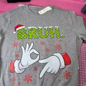 May include: Gray crew neck t-shirt with the word "BRUH" in green checkered letters, a Santa hat, and cartoon hands making the "okay" gesture. Red snowflakes are scattered around the design.