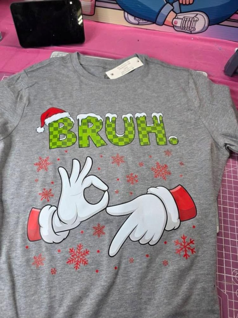 May include: Gray crew neck t-shirt with the word "BRUH" in green checkered letters, a Santa hat, and cartoon hands making the "okay" gesture. Red snowflakes are scattered around the design.