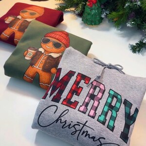 May include: Folded sweatshirts with holiday designs. One is gray with "MERRY Christmas" in plaid lettering. Another is green with a gingerbread man graphic. The third is burgundy with a gingerbread man graphic.