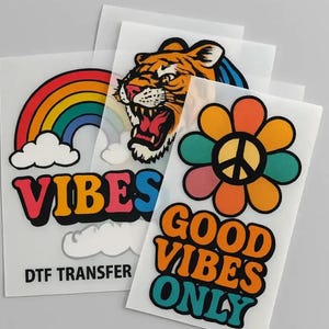 May include: A collection of DTF transfer sheets featuring colorful designs. Designs include a rainbow with the word "VIBES", a roaring tiger, and a flower with a peace sign and the words "GOOD VIBES ONLY".