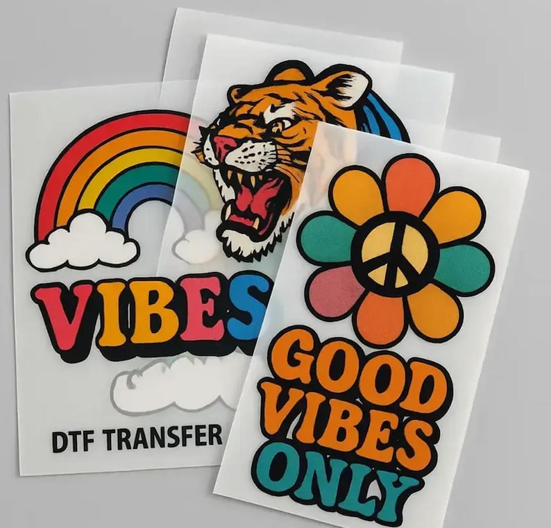 May include: A collection of DTF transfer sheets featuring colorful designs. Designs include a rainbow with the word "VIBES", a roaring tiger, and a flower with a peace sign and the words "GOOD VIBES ONLY".