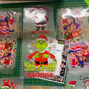 May include: A collection of Christmas-themed heat transfer vinyl designs. The designs include Santa Claus, the Grinch, and cartoon characters, with text that reads "Santa Says" and "Six Seven". The designs are on a green cutting mat.