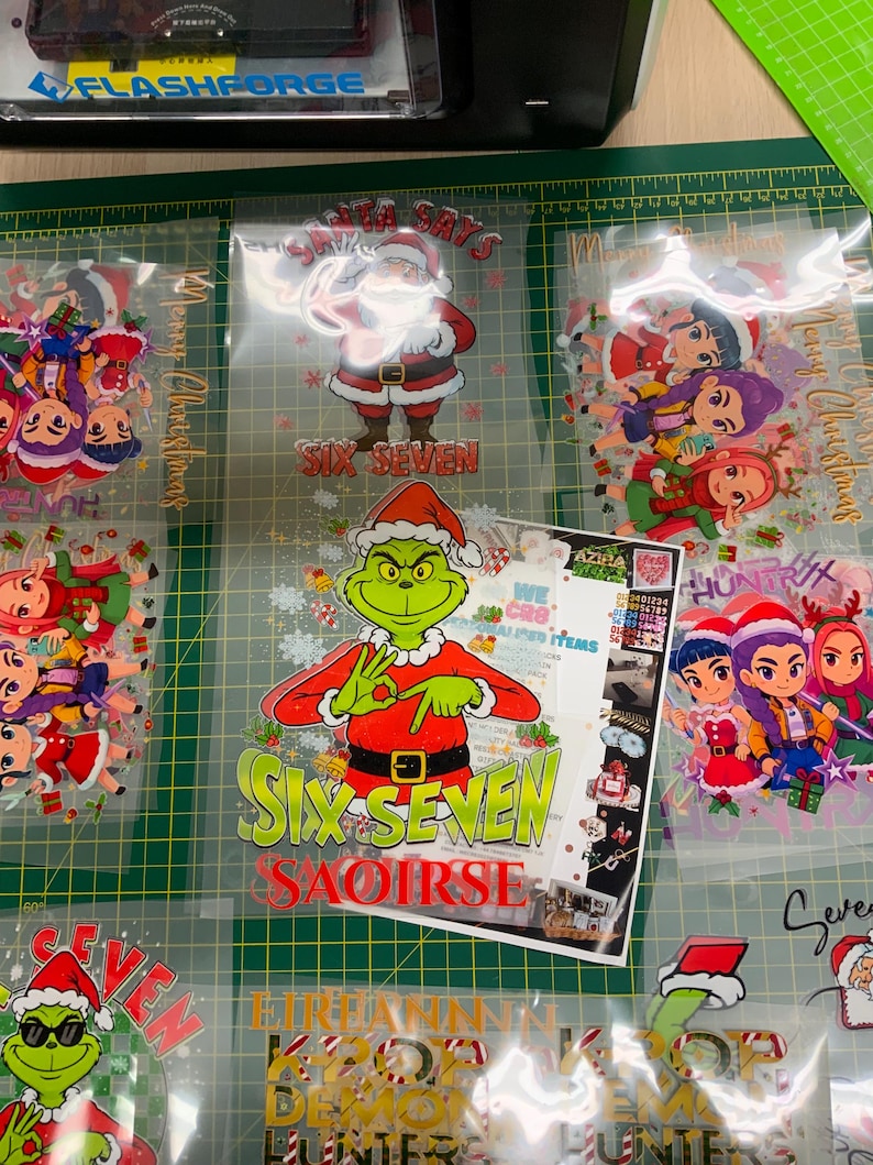 May include: A collection of Christmas-themed heat transfer vinyl designs. The designs include Santa Claus, the Grinch, and cartoon characters, with text that reads "Santa Says" and "Six Seven". The designs are on a green cutting mat.
