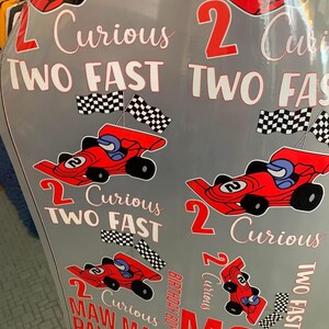 May include: A gray surface decorated with red race cars, checkered flags, and the phrases "2 Curious" and "Two Fast." The text "Birthday Boy," "Mom," and "Aunt" are also visible.