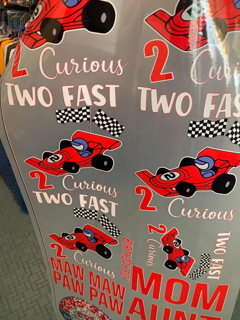 May include: A gray surface decorated with red race cars, checkered flags, and the phrases "2 Curious" and "Two Fast." The text "Birthday Boy," "Mom," and "Aunt" are also visible.
