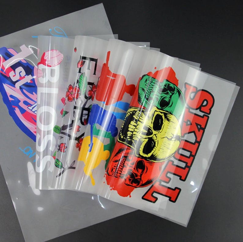 May include: A stack of transparent transfer sheets with colorful graphic designs. The designs include the word "BLOSSOM" with floral accents, the word "SKULL" with a skull illustration, and other text and graphics.