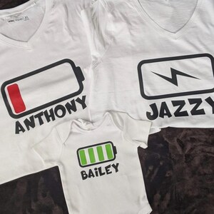 May include: White V-neck t-shirts and a baby onesie with battery graphics. The shirts have the names "Anthony" and "Jazzy" printed below the battery symbols. The onesie has the name "Bailey" printed below a green battery graphic.