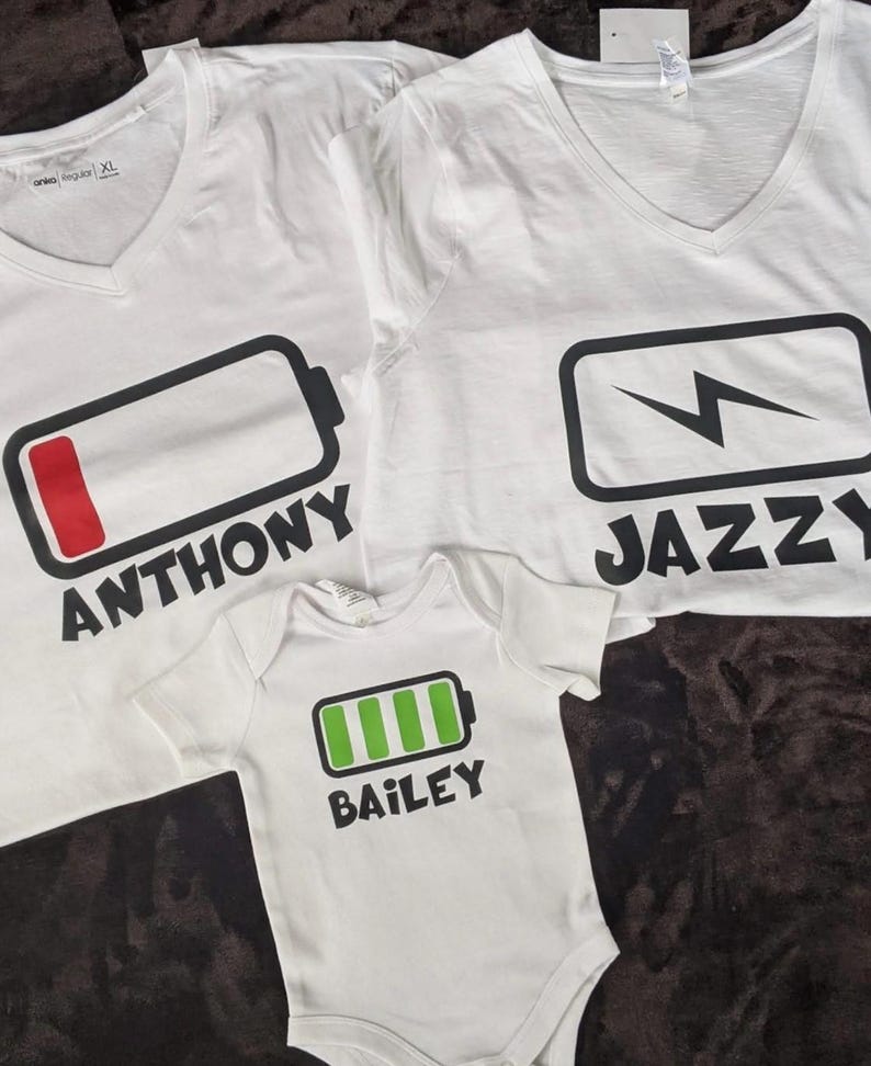 May include: White V-neck t-shirts and a baby onesie with battery graphics. The shirts have the names "Anthony" and "Jazzy" printed below the battery symbols. The onesie has the name "Bailey" printed below a green battery graphic.