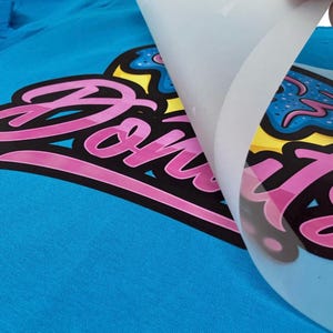 May include: A close-up of a blue t-shirt with a pink and black graphic that says "Donuts". The graphic is partially covered by a clear sheet. The graphic also includes a yellow and blue donut design.