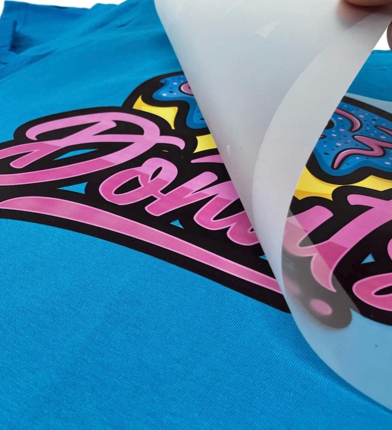 May include: A close-up of a blue t-shirt with a pink and black graphic that says "Donuts". The graphic is partially covered by a clear sheet. The graphic also includes a yellow and blue donut design.