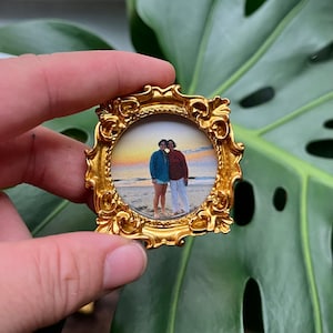 May include: A gold-colored ornate frame with a photo of two people standing on a beach. The photo is in color and shows a sunset in the background.