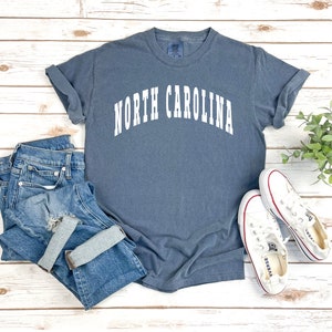 Pu&ograve; includere: Una maglietta blu con la scritta "NORTH CAROLINA" in lettere bianche. La maglietta &egrave; stesa su una superficie di legno bianco con un paio di scarpe di tela bianche e un paio di jeans blu.