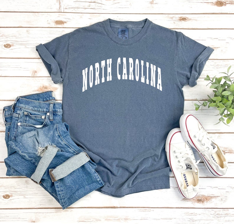 Pu&ograve; includere: Una maglietta blu con la scritta "NORTH CAROLINA" in lettere bianche. La maglietta &egrave; stesa su una superficie di legno bianco con un paio di scarpe di tela bianche e un paio di jeans blu.