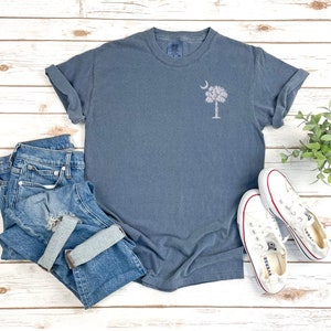 May include: A blue t-shirt with a white embroidered image of a palmetto tree and crescent moon. The t-shirt is laid flat on a white wooden surface with a pair of white canvas shoes and blue jeans.
