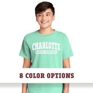 May include: A light green t-shirt with a white graphic that says "CHARLOTTE NORTH CAROLINA" in a vintage collegiate style font.