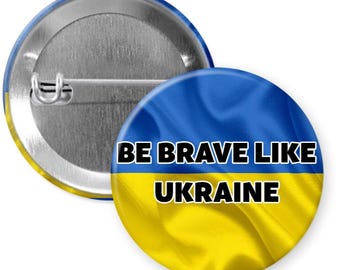 May include: A blue and yellow button with the text "BE BRAVE LIKE UKRAINE" on it. The button is a round pinback button with a metal back.