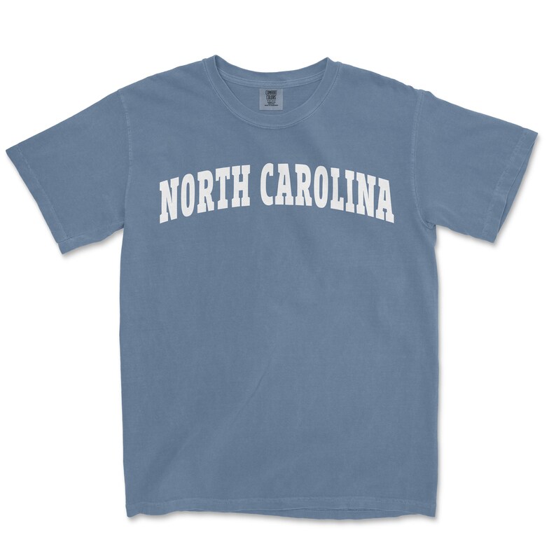 Pu&ograve; includere: Una maglietta blu chiaro con il testo "NORTH CAROLINA" in bianco.